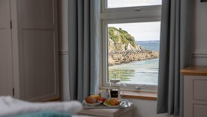 Une chambre à coucher avec un lit, une table de chevet sur laquelle se trouve un plateau avec des articles pour le petit-déjeuner, et une fenêtre donnant sur un paysage côtier.