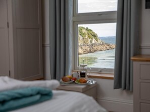 Ein Schlafzimmer mit einem Bett, einem Nachttisch mit Frühstücksgedecken und einem Fenster mit Blick auf eine Küstenlandschaft.