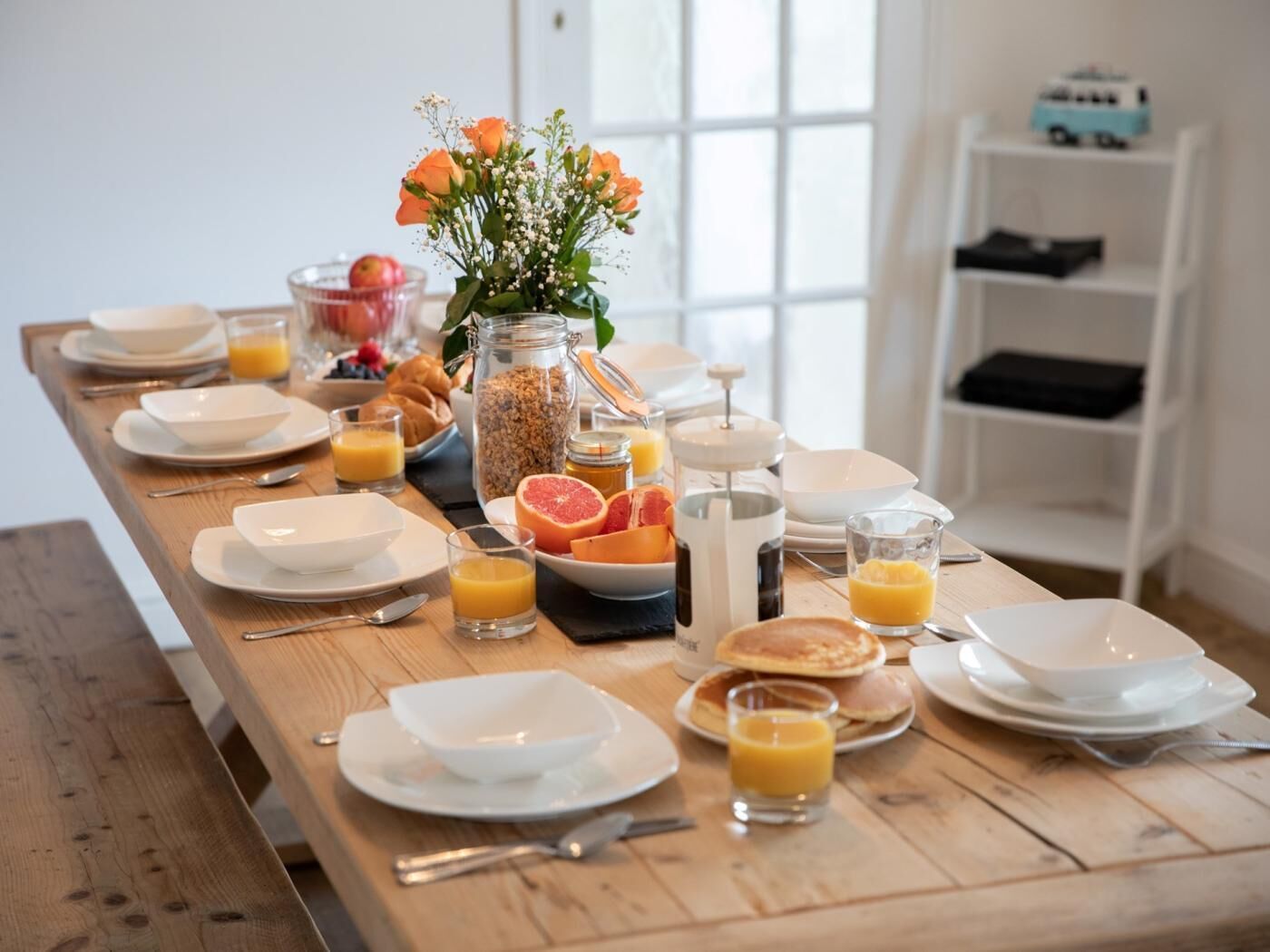 Una tavola imbandita per la colazione con piatti, ciotole, bicchieri e una coppetta di frutta.