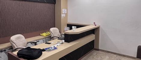 A reception desk with a computer, a telephone, and a bag on it. There is a chair and a desk lamp.