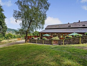 Un bâtiment en pierre avec un toit en tuiles, entouré d’un jardin clos avec des plantes en pot et une clôture en bois.