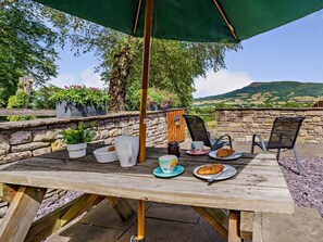 Une table en bois, un parasol vert, des plantes en pot et une vue panoramique sur des collines.
