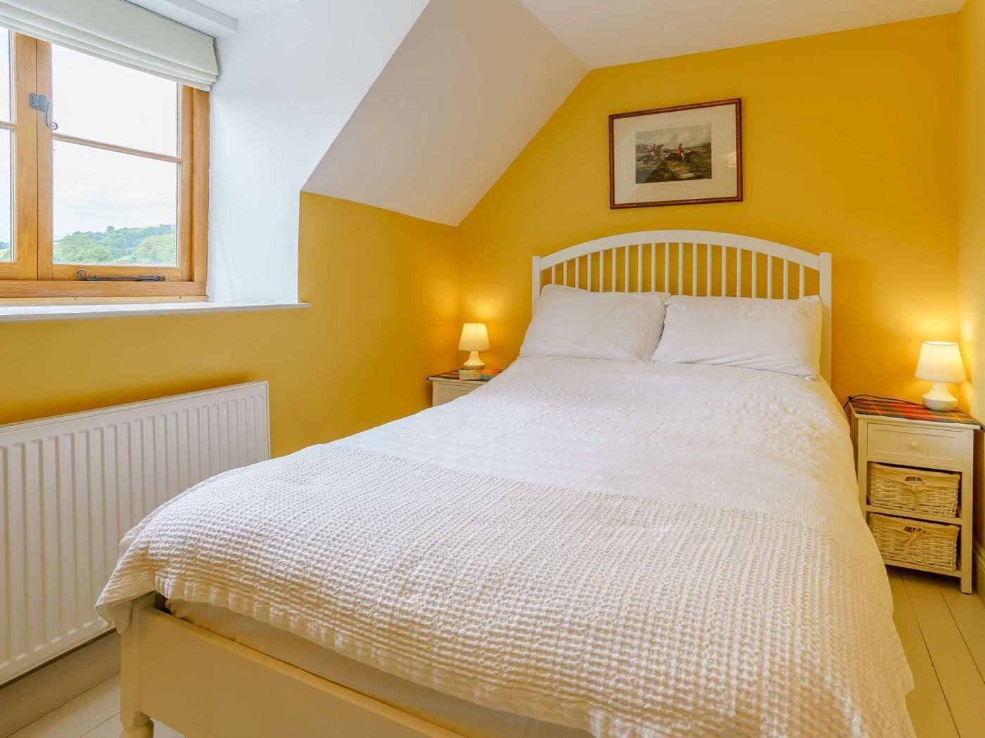 Quarto com cama, duas luminárias de cabeceira, janela, radiador e um quadro na parede.