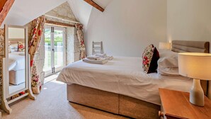 A bedroom with a bed, a nightstand, a lamp, a mirror, and a view of a garden through a window. - 3 Bed in Shaftesbury (oc-48804) (East Melbury)