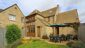 Une maison en pierre avec une grande fenêtre, une porte en bois et une terrasse équipée de chaises et d’un parasol.