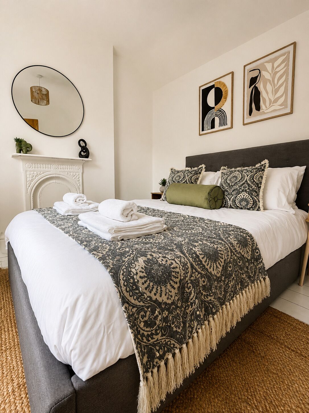 A neatly made bed with a patterned throw, a round mirror, and two framed artworks on the wall.