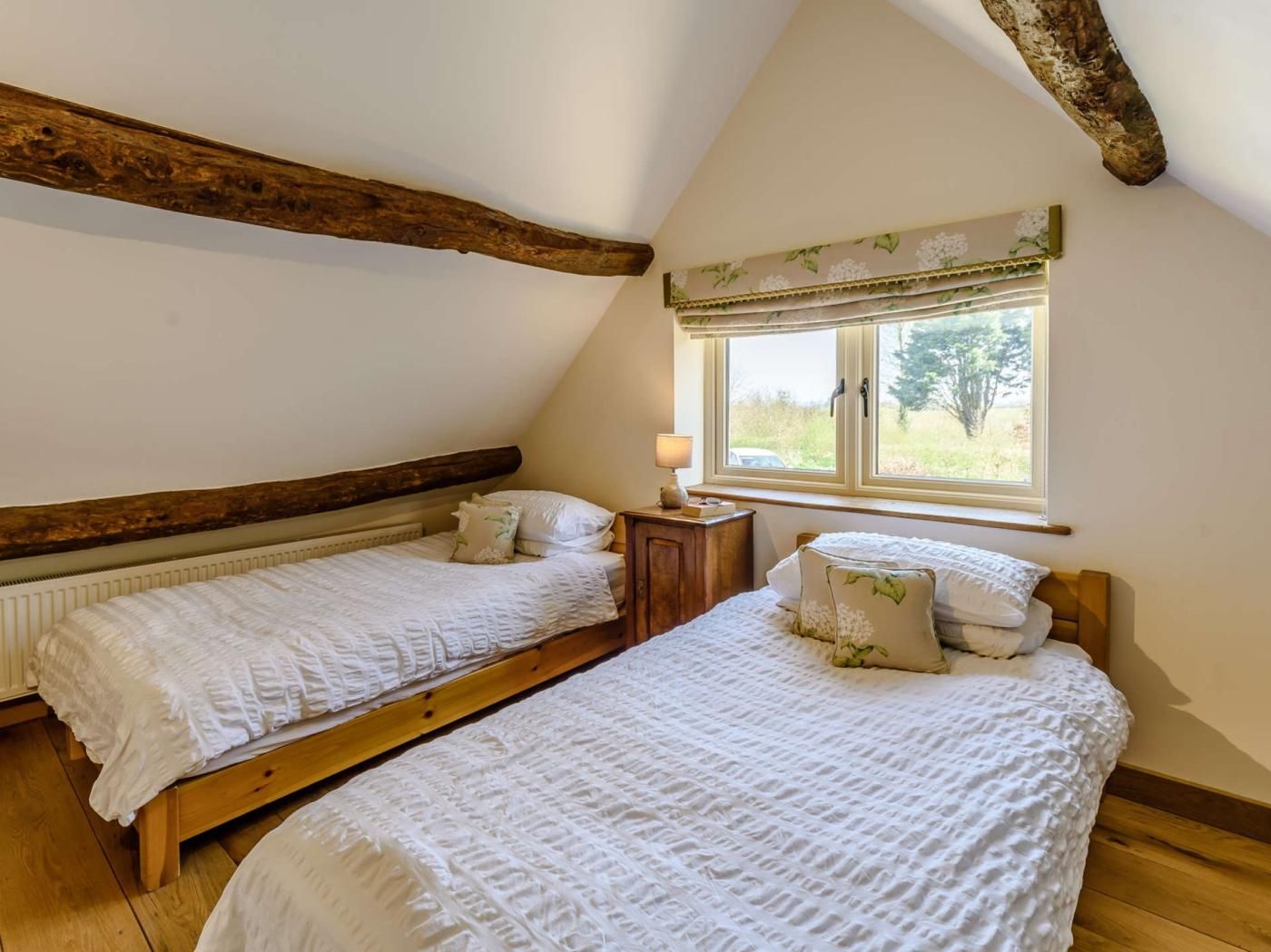 A bedroom with two beds, wooden beams, a window with a view, and a bedside table with a lamp.