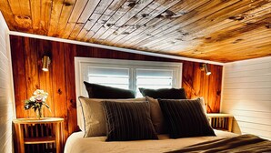 A bedroom with a wooden ceiling, a bed with multiple pillows, a nightstand with a vase of flowers, and a window with blinds.