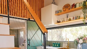 A modern kitchen with a wooden staircase, a dining area with stools, and a view through a window.