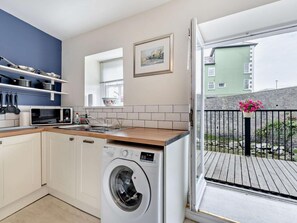 A compact kitchen with a washing machine, a microwave, and a window with a view of a building. - 1 Bed in Aberaeron (oc-49261) (Aberaeron)