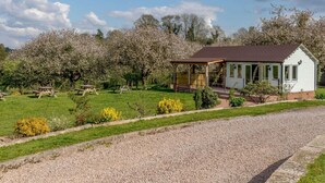 Een wit huisje met een veranda, omringd door een gazon met picknicktafels en bloeiende bomen.