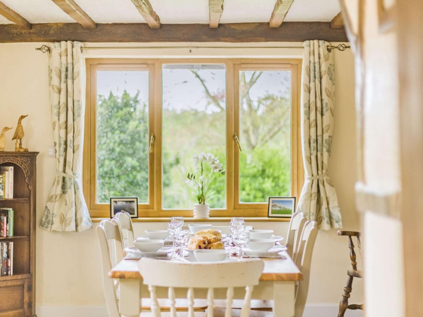 A dining table set with plates, glasses, and a centerpiece, positioned in front of a window with curtains.