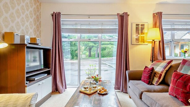 A living room with a wooden coffee table, a sofa, a television, and a window with curtains. - 2 Bed in Cheltenham (oc-49336) (Woodmancote)