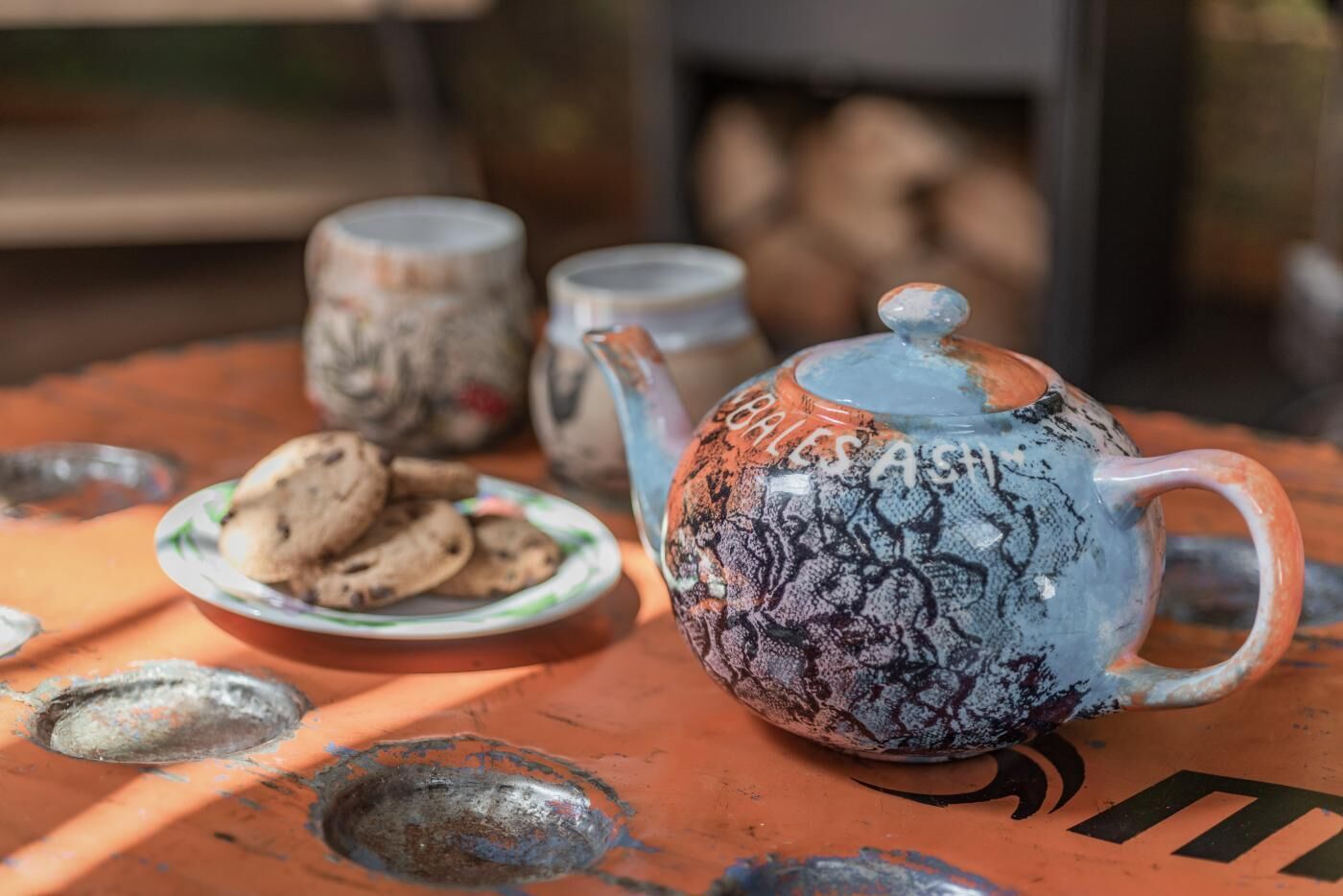 A teapot with a floral pattern, a plate with cookies, and two cups on a table with a rustic appearance.