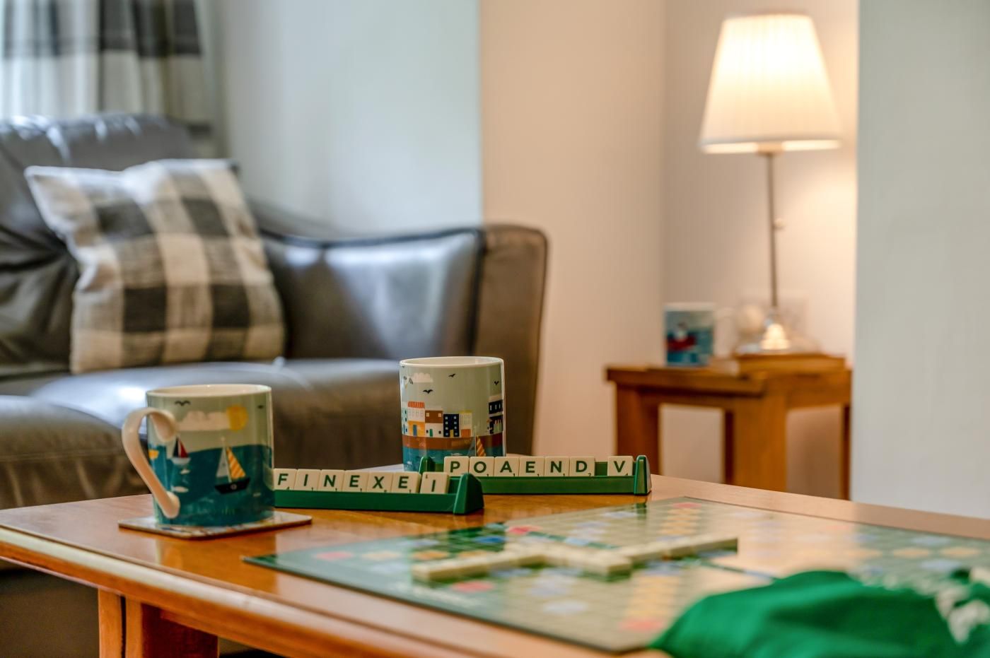 A cozy living room with a sofa, a coffee table with a Scrabble game, and two mugs with nautical designs.