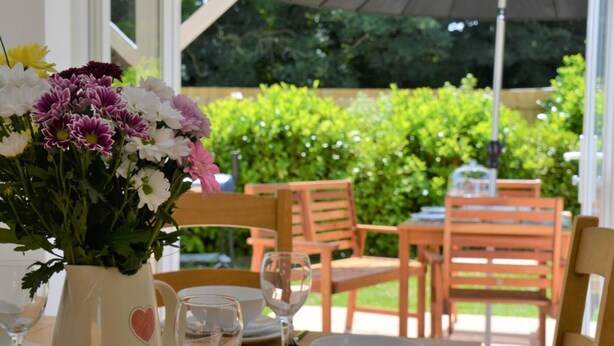 Un'area pranzo all'aperto con un tavolo apparecchiato per due persone, un vaso di fiori e una sedia in legno.