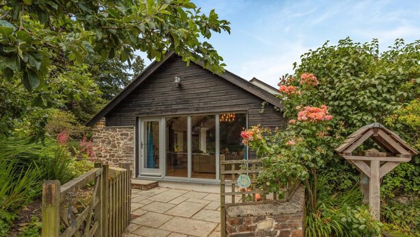 A rustic wooden cottage with a stone exterior, a wooden gate, and a garden with flowering plants. - 1 Bed in Umberleigh (oc-49554) (High Bickington)