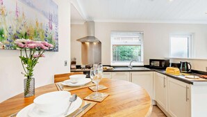 Een moderne keuken met witte kastjes, een houten eettafel gedekt voor twee personen, een schilderij aan de muur en een raam met jaloezieën.