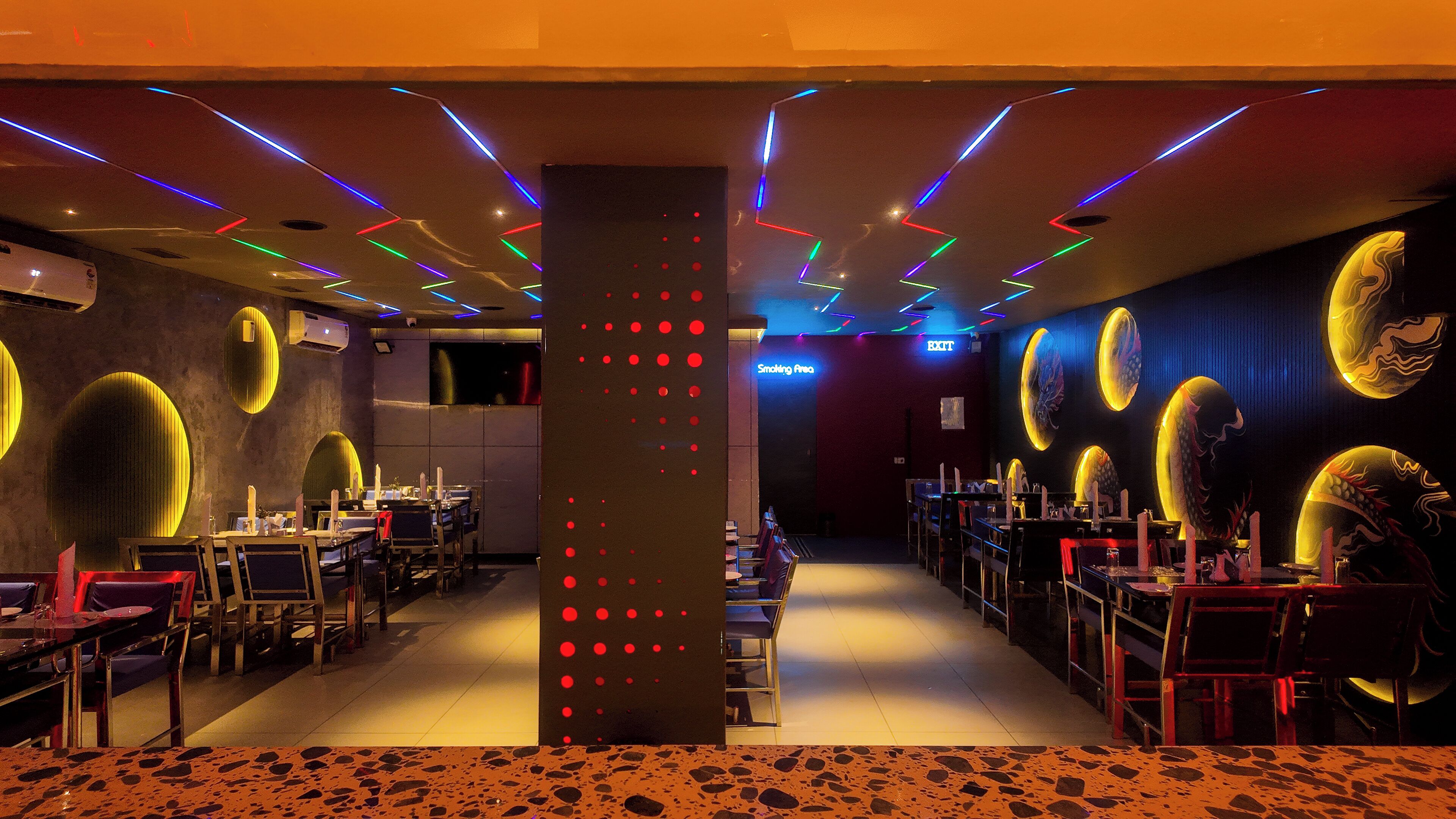 Interior de restaurante con una columna central, mesas, sillas y un letrero de neón que indica zona para fumadores.