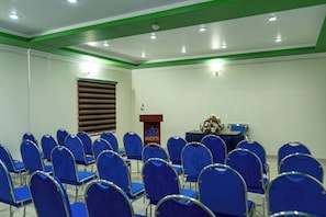 Un salón de conferencias con filas de sillas azules, un atril con un logotipo y una ventana con persianas.