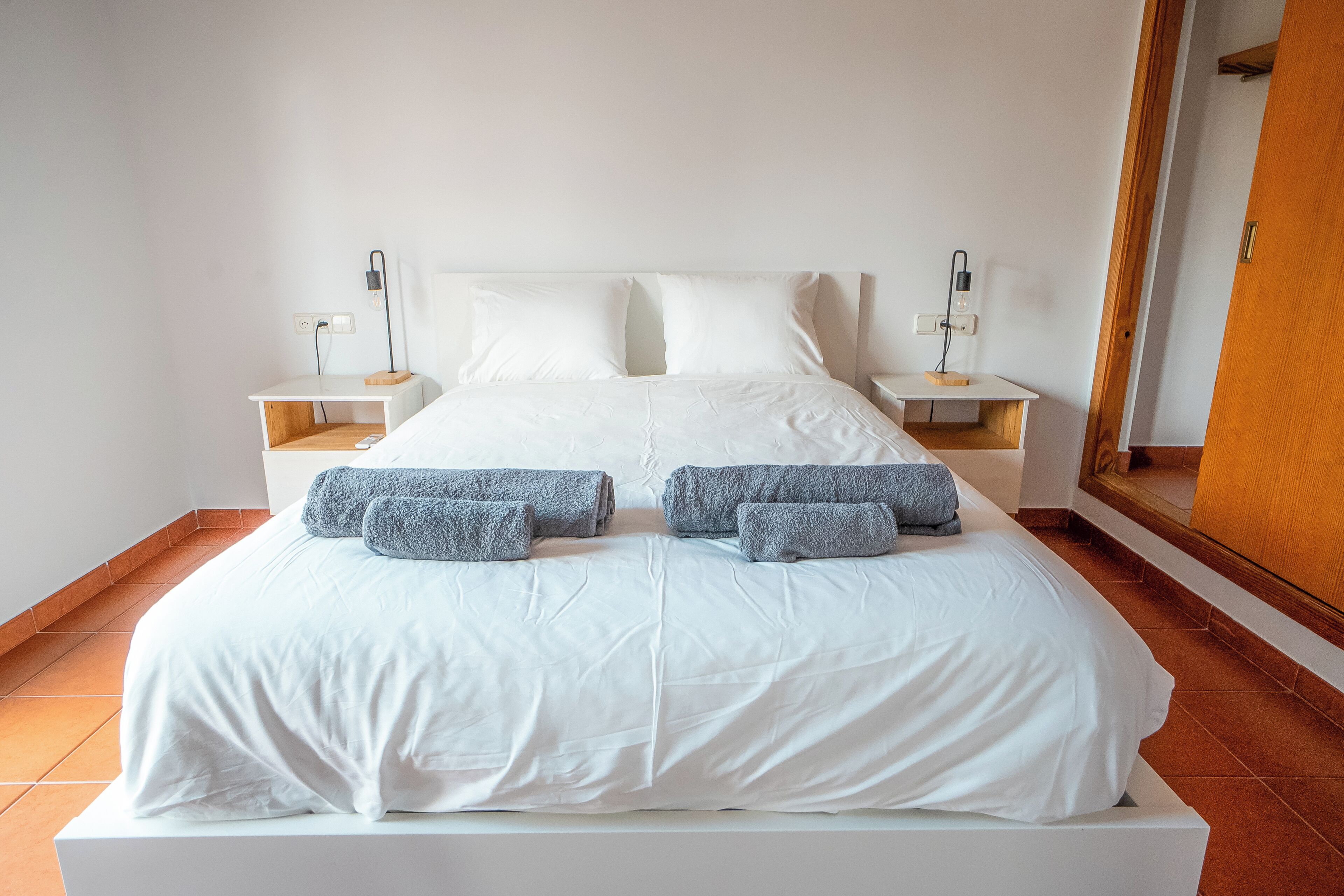 Un lit bien fait, avec du linge de lit blanc et des serviettes grises, deux tables de chevet avec des lampes, et une porte en bois à l’arrière-plan.