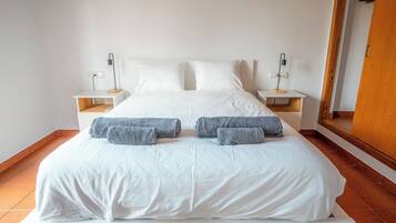 Un lit bien fait, avec du linge de lit blanc et des serviettes grises, deux tables de chevet avec des lampes, et une porte en bois à l’arrière-plan.