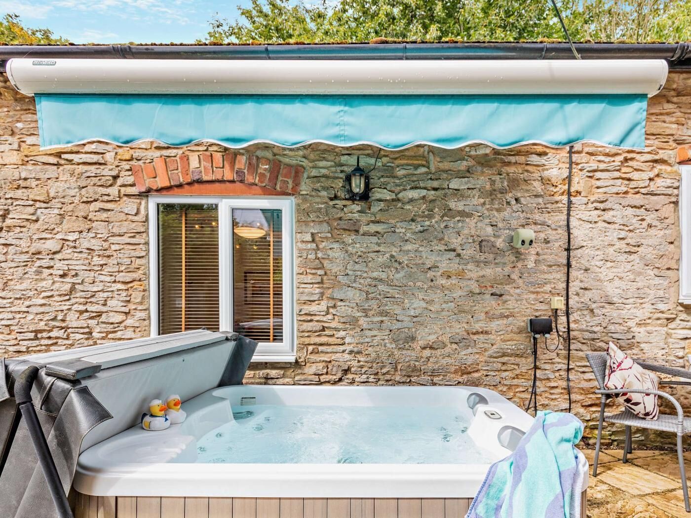 A stone building with a hot tub, a window with blinds, and a chair.