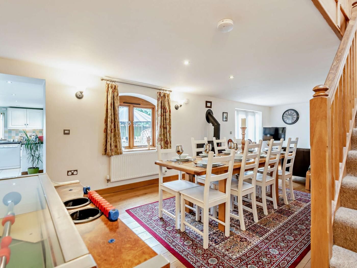 A dining area with a wooden table and white chairs, a kitchen with white cabinets, a radiator, and a window with curtains.
