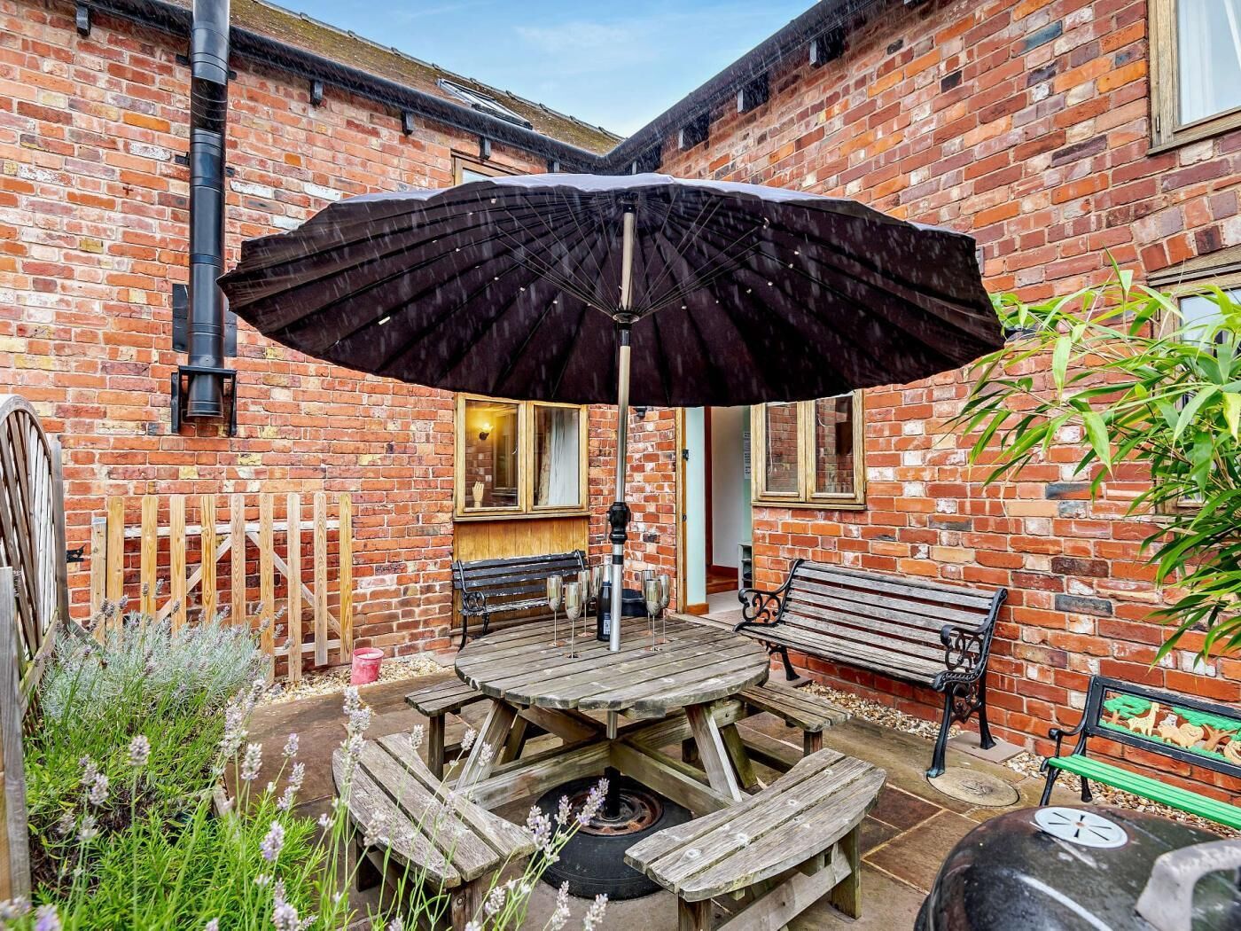 A brick building with a wooden table and benches, a black umbrella, and a grill.