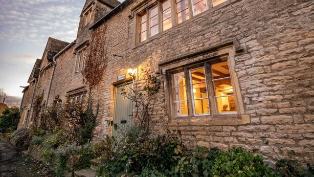 Ein Gebäude aus Stein mit mehreren Fenstern, von denen einige mit Vorhängen versehen sind, und einem kleinen Garten mit Pflanzen und Sträuchern.