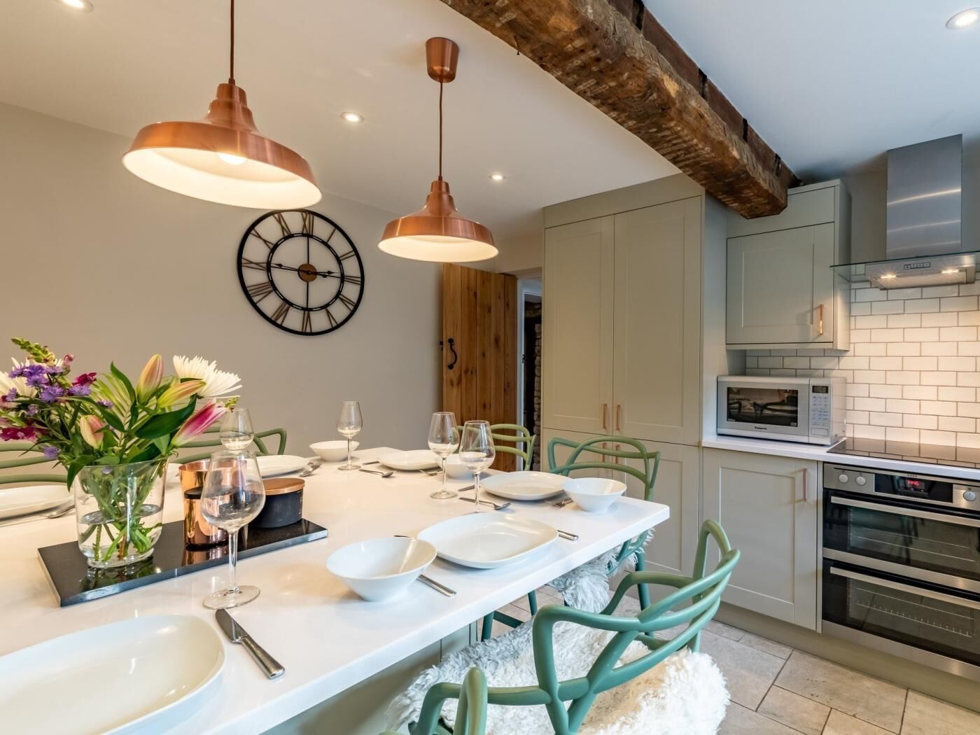 A modern kitchen with a dining table set for six, featuring green chairs, pendant lighting, and a clock on the wall.