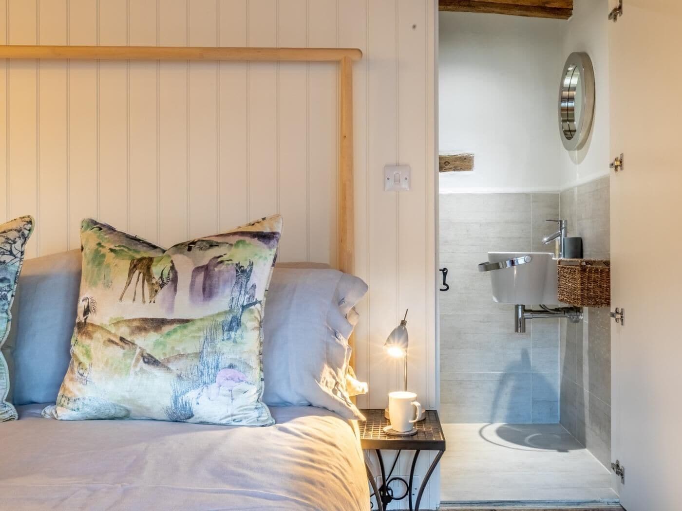 A bedroom with a bed, pillows, bedside table, lamp, and a view into a bathroom with a sink and mirror.