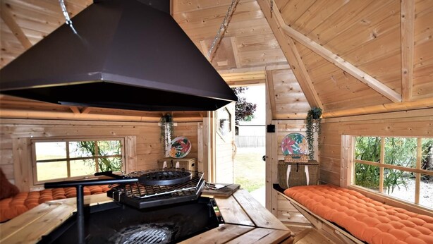 A wooden cabin interior with a central fireplace, a bed with an orange mattress, and a window with a view of greenery. - 4 Bed in Umberleigh (oc-50154) (Burrington)