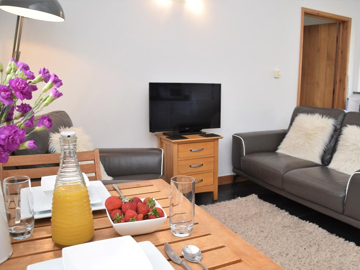 A living room with a sofa, a television, a wooden table with a bowl of strawberries, a bottle of juice, and a vase of flowers.