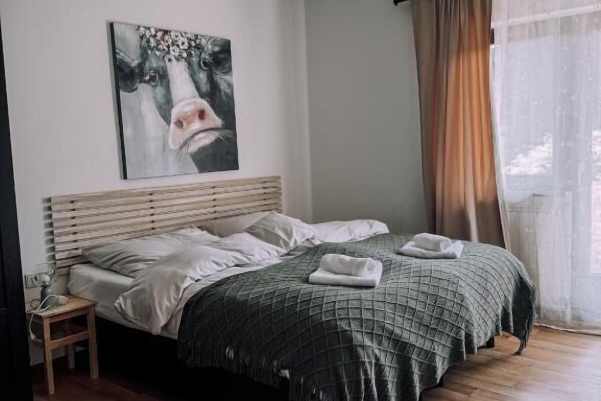 A bedroom with a bed, a bedside table, a hanging light fixture, and a window with curtains.