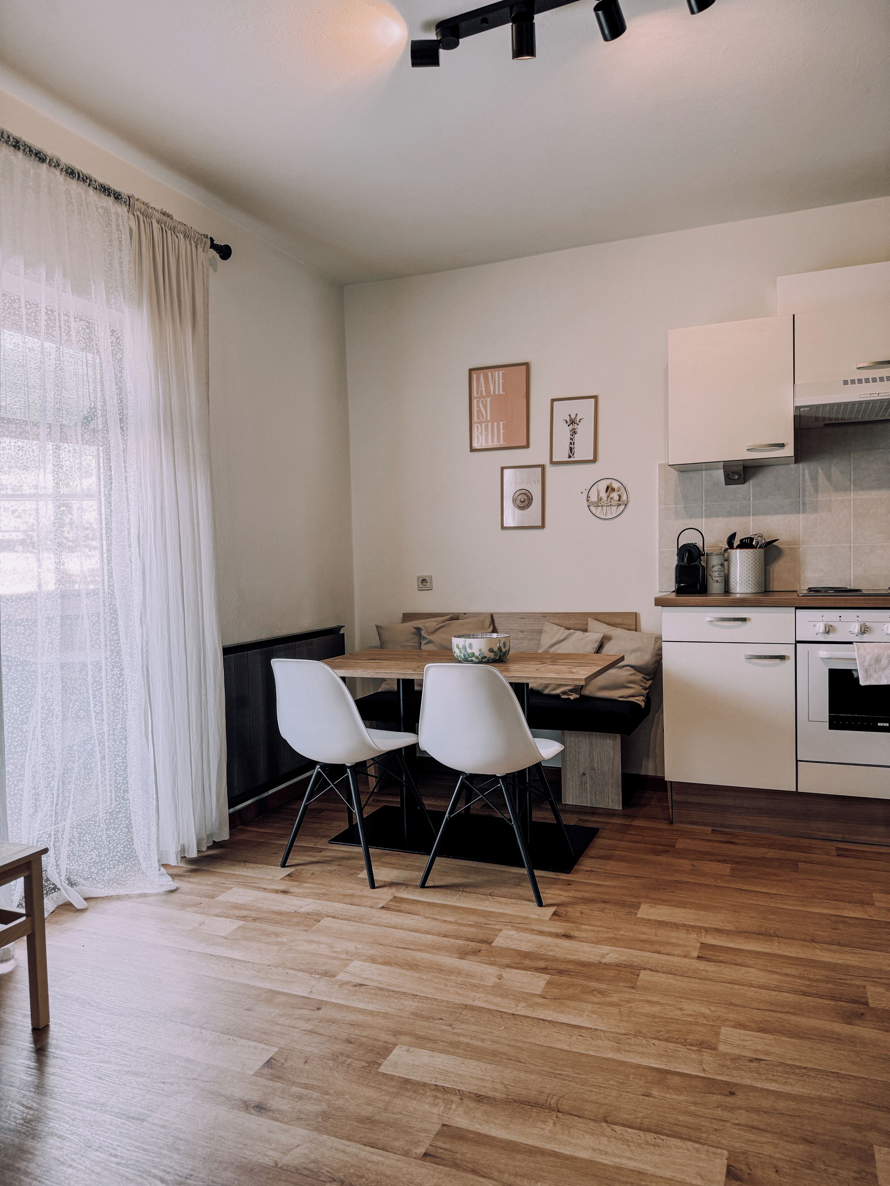 A modern kitchen with white cabinets, a dining area with two white chairs, and a wooden dining table. There are framed pictures on the wall and a window with sheer curtains.