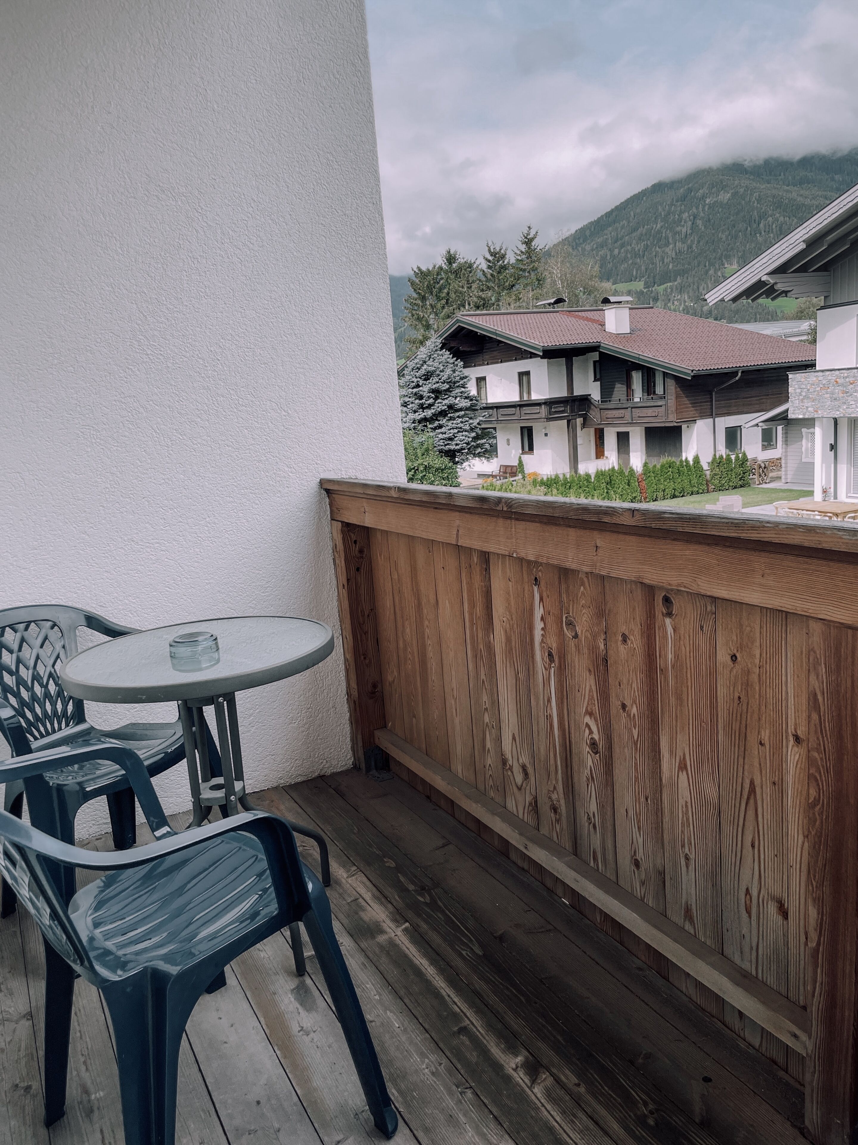 A wooden deck with a round table and chairs, a white wall, and a view of houses and mountains in the background.