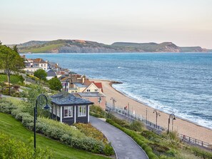 A coastal town with a paved walkway leading to a beach, houses with visible windows, and a clear view of the sea.