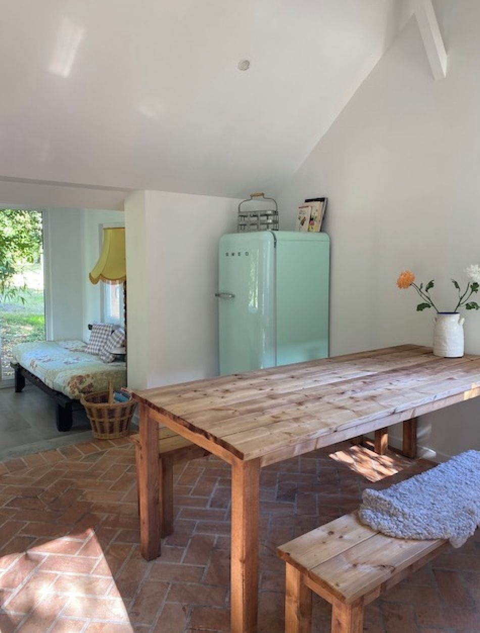 A room with a wooden table, a bench, a refrigerator, and a window with a view of greenery.