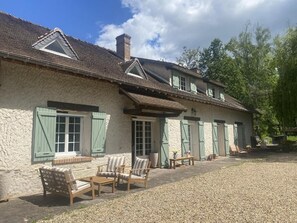 Un edificio de piedra con un patio de grava, muebles de madera y contraventanas verdes.