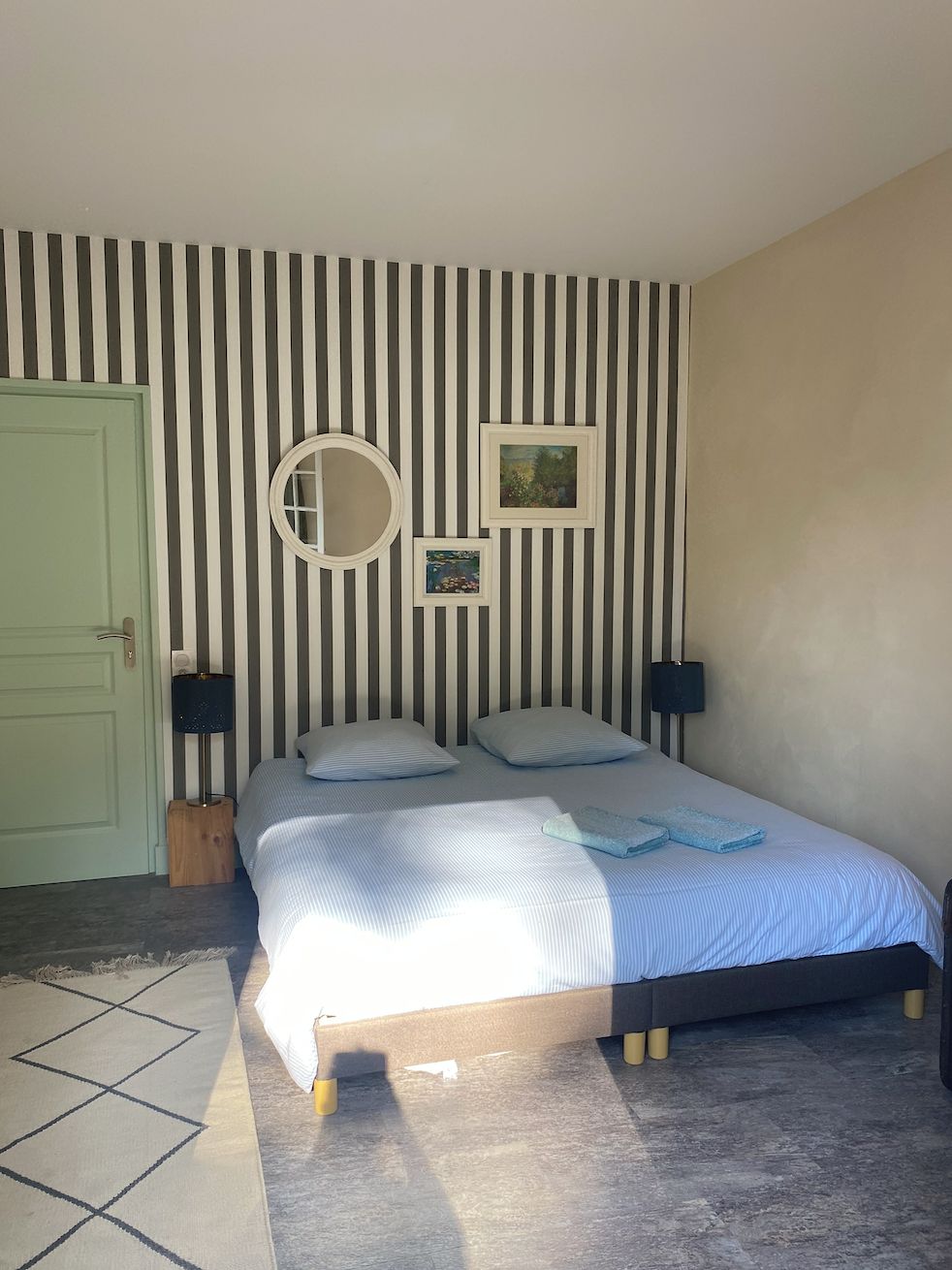 Un dormitorio con cama, una puerta, una pared con rayas verticales, un espejo redondo y dos cuadros enmarcados.