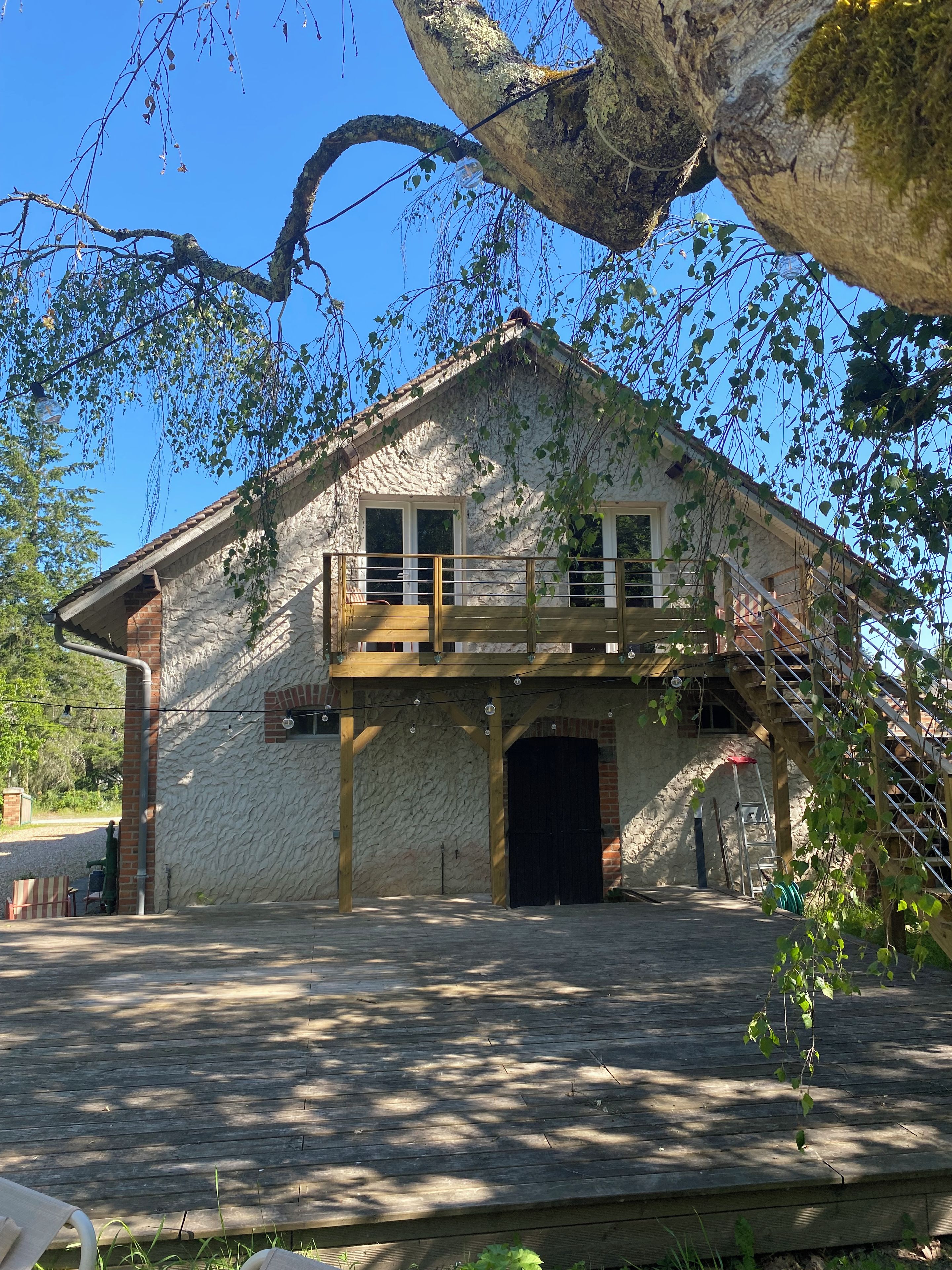 Un edificio rustico in pietra con un balcone in legno, un grande albero in primo piano e un cielo azzurro limpido.