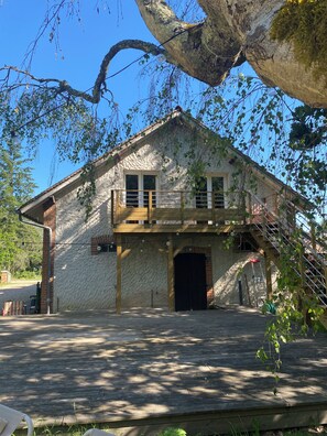 Un edificio rústico de piedra con un balcón de madera, un árbol grande en primer plano y un cielo azul despejado.