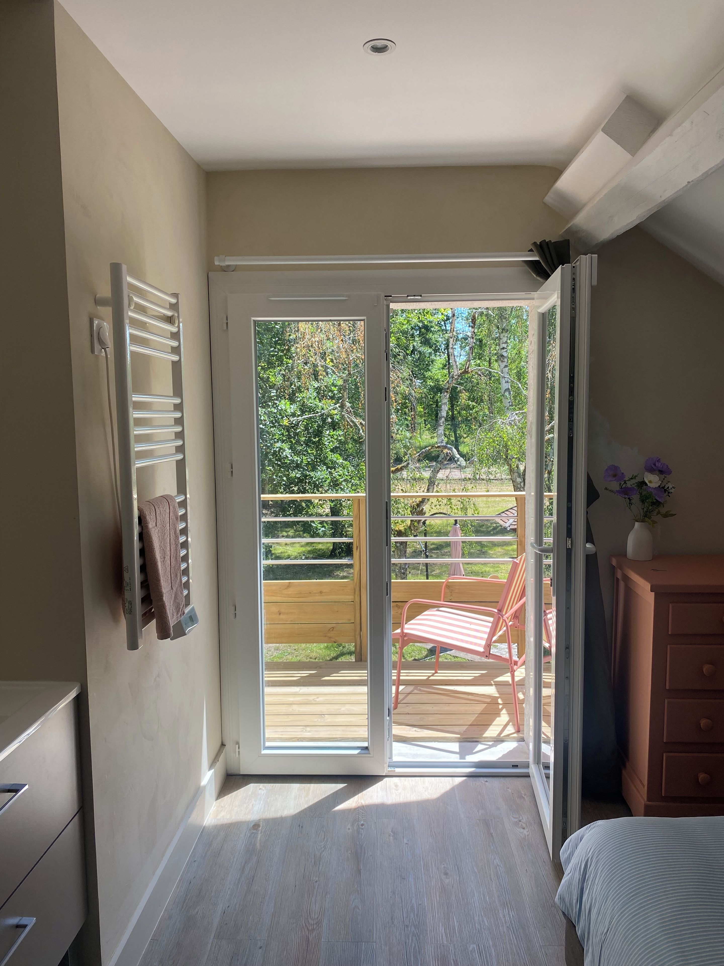 A room with a sliding glass door leading to an outdoor deck, a wooden chair, and a dresser with a vase of flowers.
