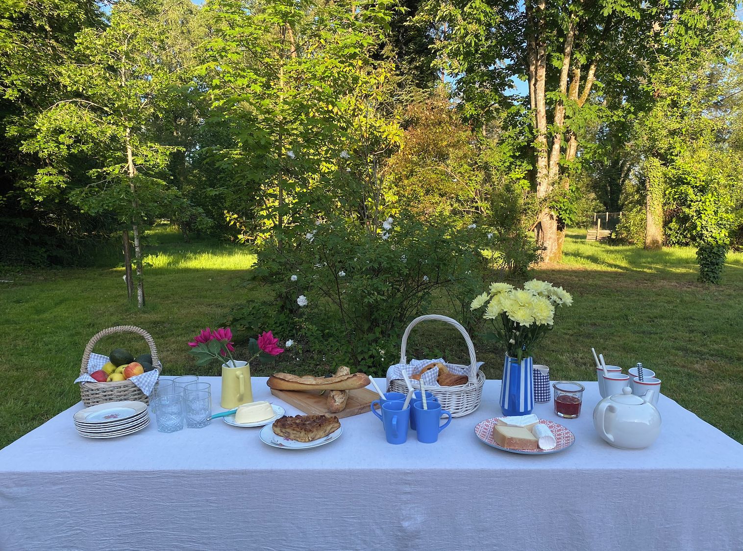 An outdoor picnic setup with a table set for two, including plates, glasses, a basket of fruit, a loaf of bread, and a vase of flowers.