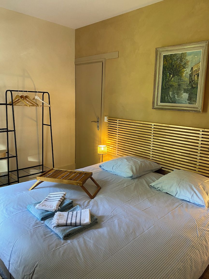 A neatly made bed with striped bedding and two pillows, a wooden headboard, a painting on the wall, and a hanging rack with clothes.