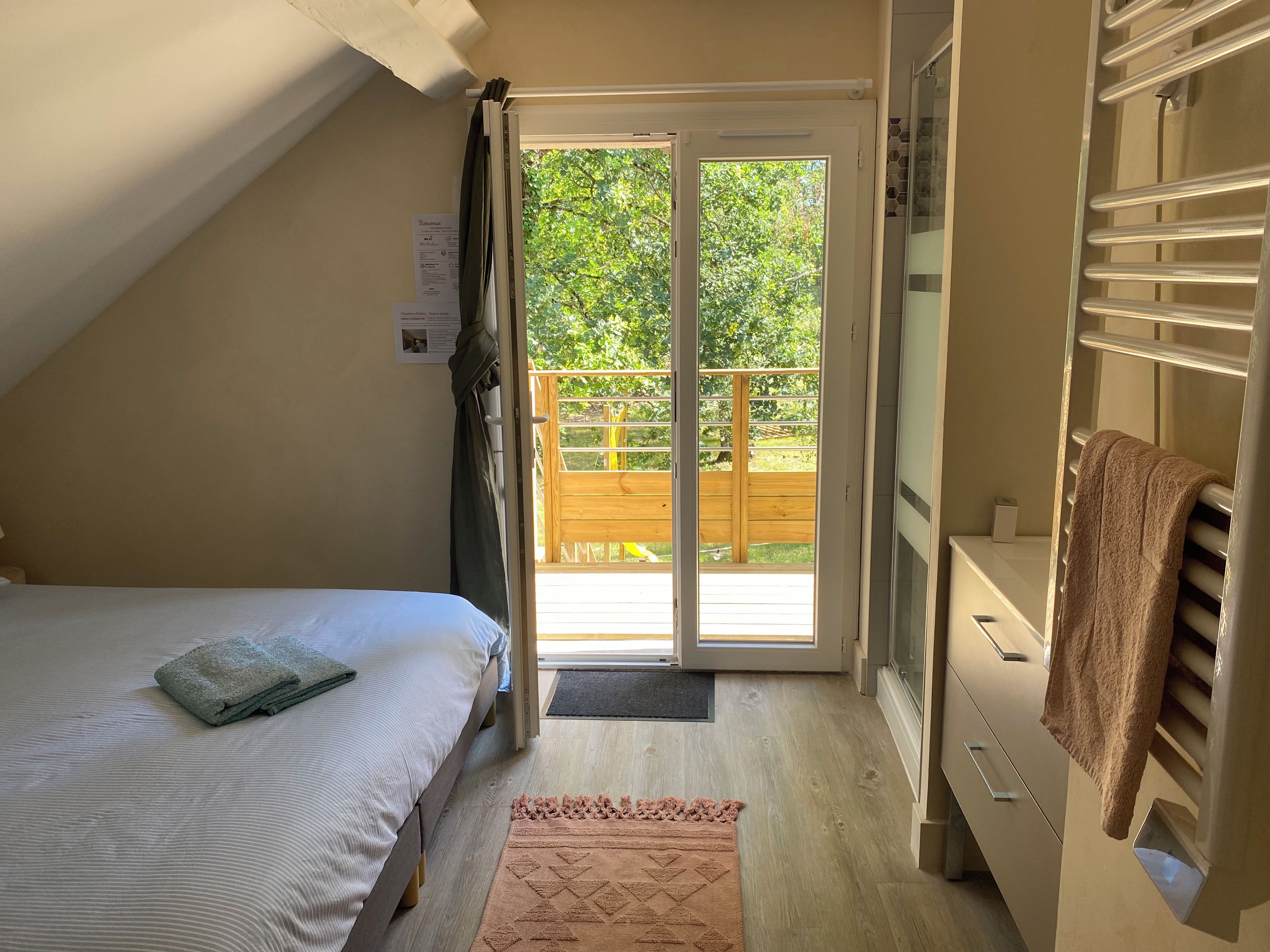 Un dormitorio con cama, piso de madera, puerta de cristal corredera que da a un balcón y un perchero para toallas.