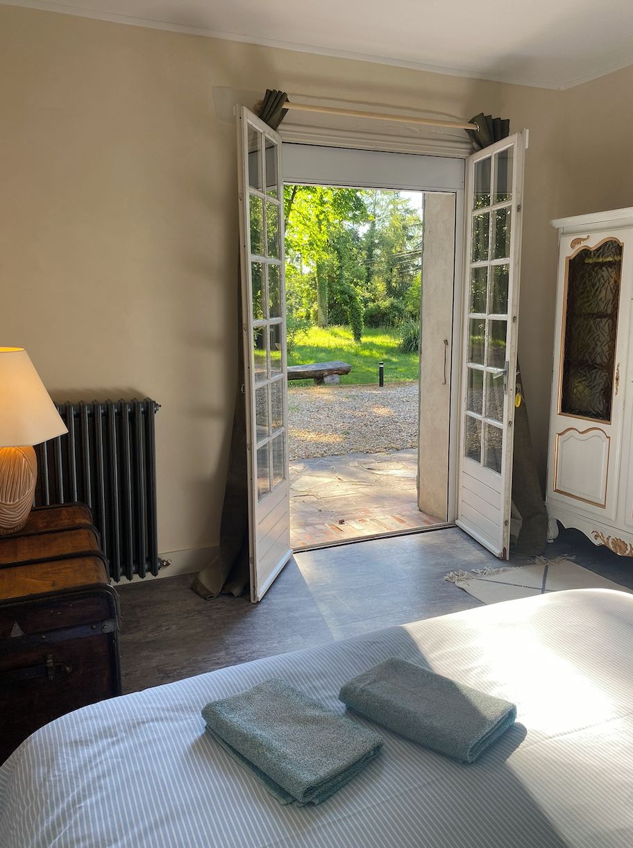 A bedroom with a bed, a radiator, a lamp, and an open door leading to an outdoor area with a bench and greenery.