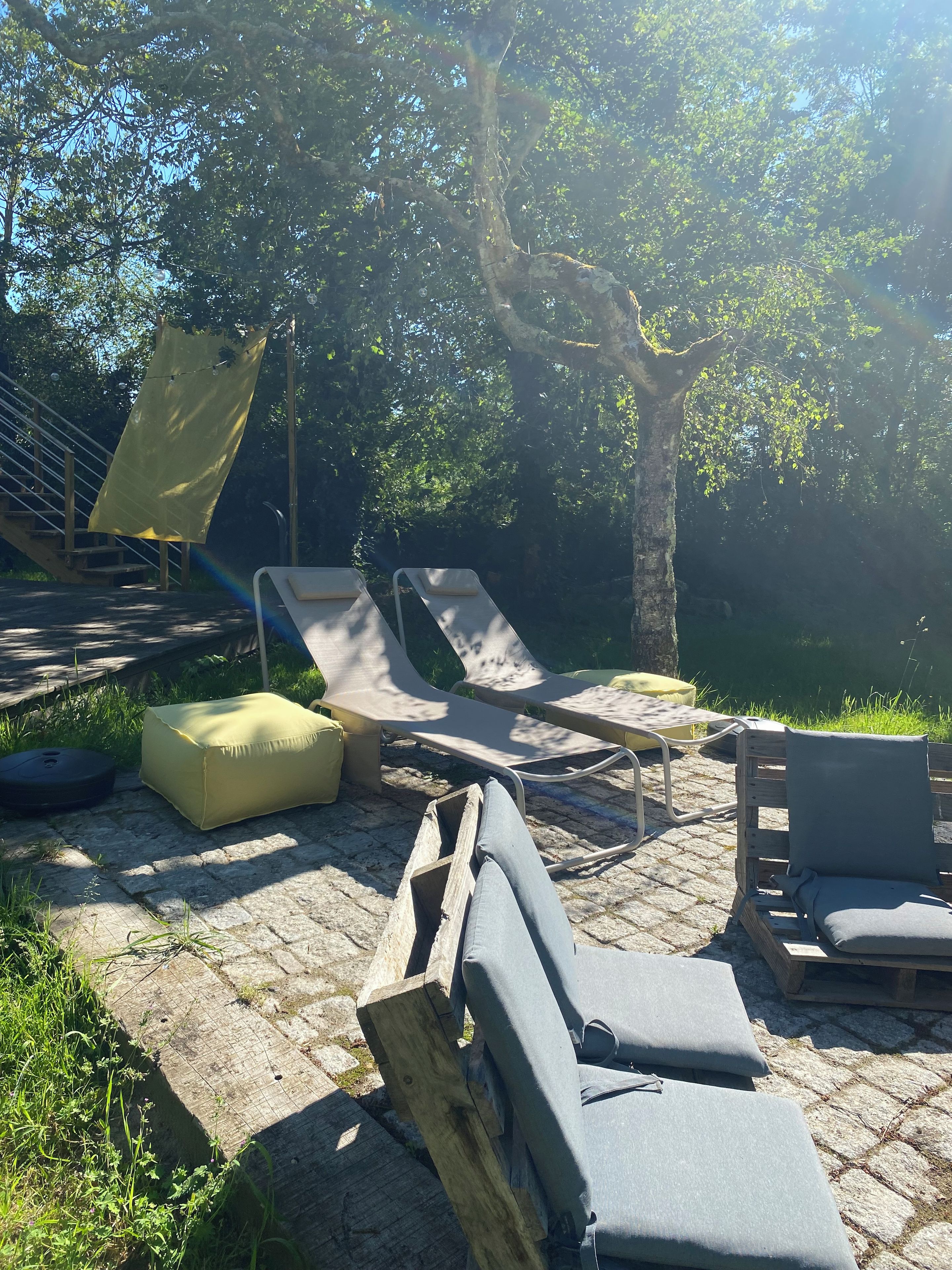 Outdoor lounge chairs, a wooden bench, and a yellow cushion on a stone patio surrounded by trees.