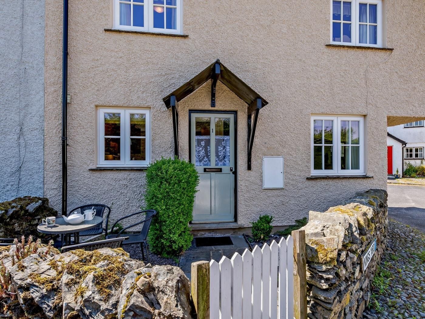 A house with a stone wall, a white picket fence, a door with a glass panel, and a small garden with a table and chairs.
